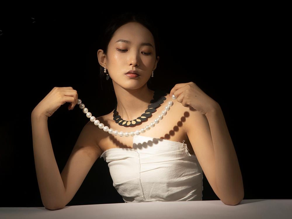 A woman holds a pearl necklace up as though she is about to put it on. She wears a dark necklace that takes the form of the abstracted shadow of the beads. 