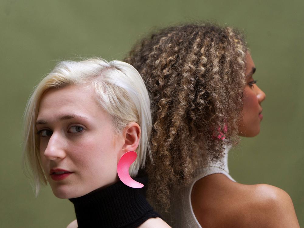 Two models stand back to back. One with curly hair and black skin faces away, one with plond hair and pale skin faces towards us.  Both wearing bright pink joyful earrings. 
