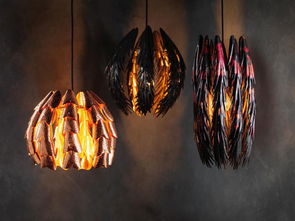 A series of 3 lighting sculptures hang against a grey textured background.   Made of organic units with hole punched textures.  Light peeks through the holes.  Each sculpture is slightly different in colour and form, from bright copper to dark blackened metal to red patinated copper forms. 