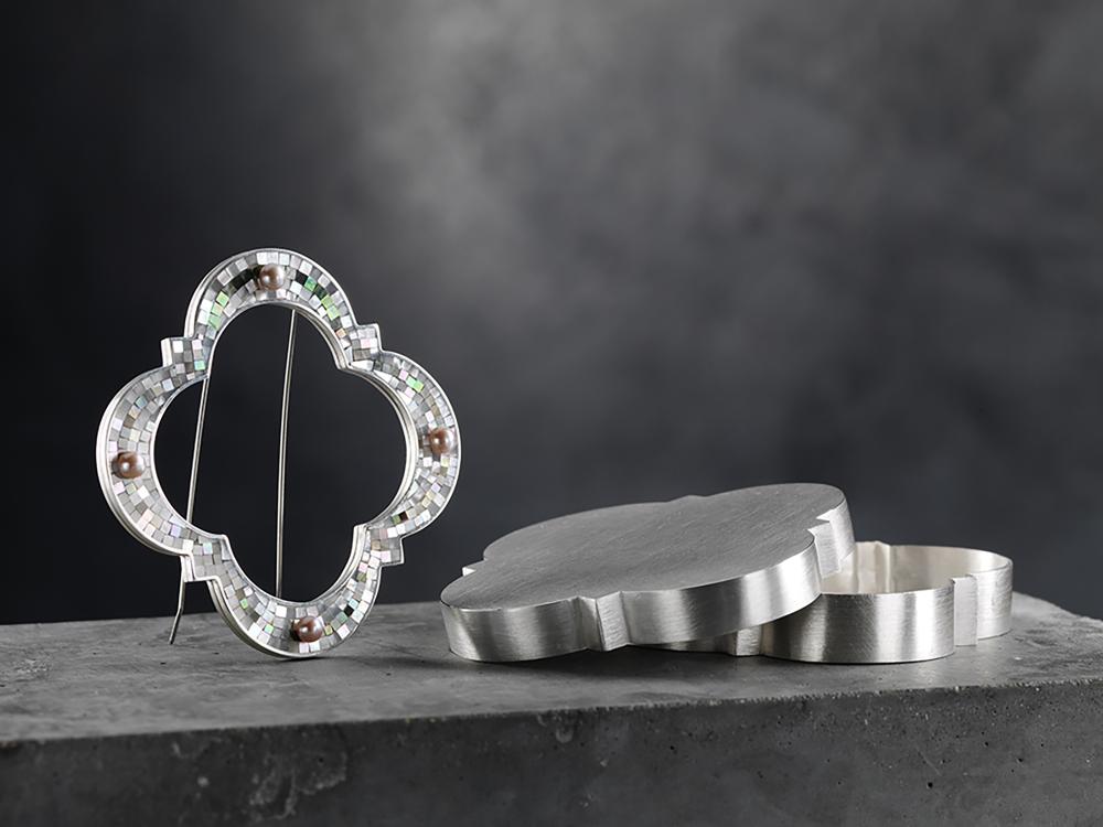 A brooch and a box rest on a concrete plinth. The brooch sits upright, showing a decorative open shape with a micro mosaic surface and 4 pearls. The box lies part opened, its form perfect for the brooch to rest within. 