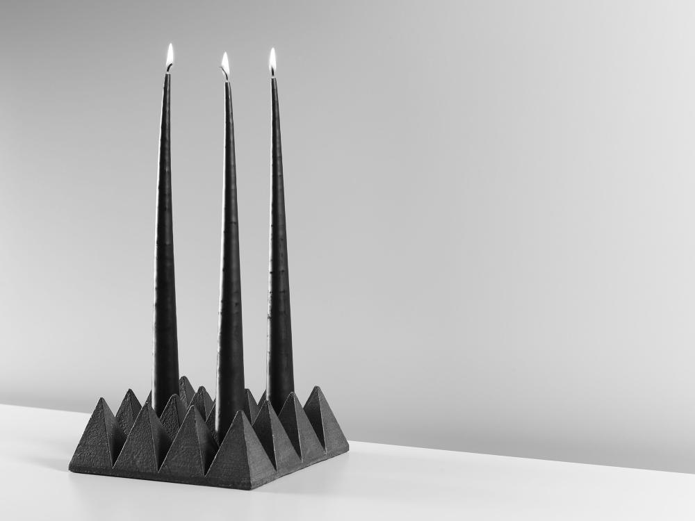 A chunky cast steel sculptural object rests on the table. It  is made of a series of triangular pyramid forms arranged in rows. Three candles are set within. 