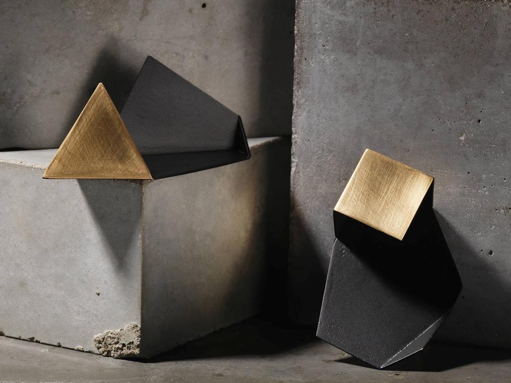 Two angular geometric earrings in black and gold rest against a concrete block. They have sharp, crisp folds and matt surface. 