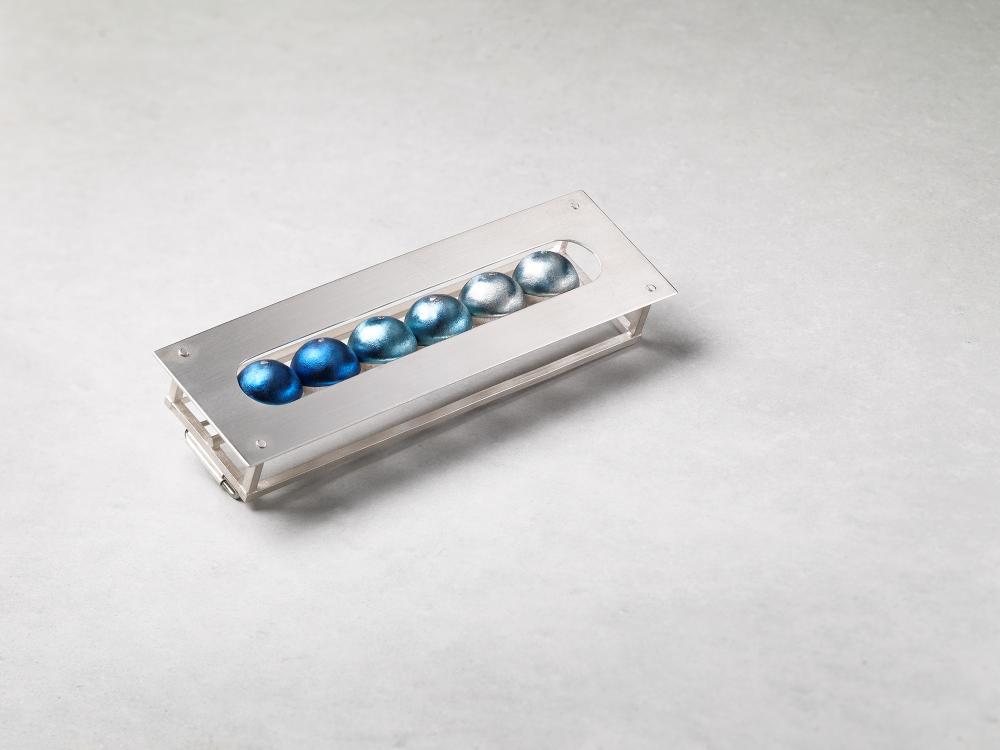 A silver brooch in a rectangular frame form rests on a grey background. 6 titanium balls sit within the brooch structure, arranged in a colour gradient from dark to light blue. 