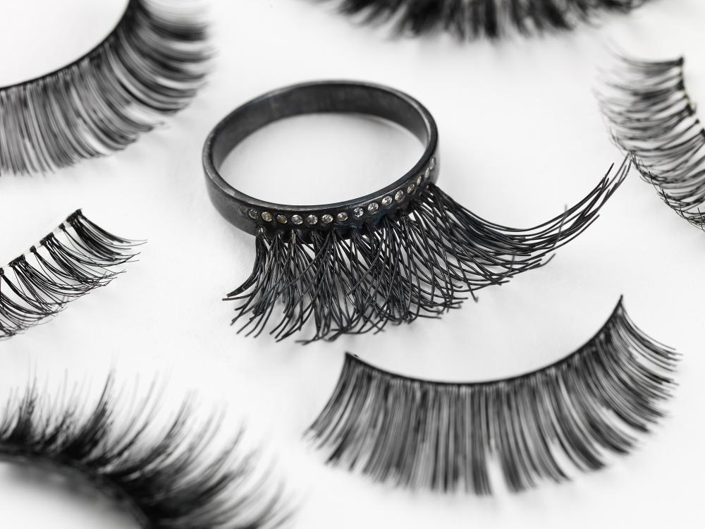 A ring sits amongst a scatter of false eyelashes. The ring is black and looks at first glance like a false eyelash. A line of tiny white stones follow the lash line. 