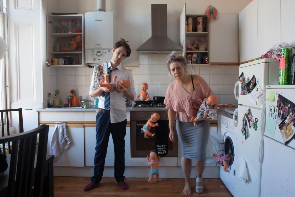 a man and a woman appearing disheveled in a kitchen, looking directly at the camera, each holding a toy baby in their arms and with multiple other toy babies scattered around the kitchen