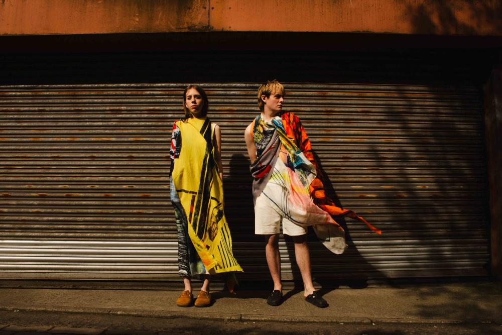 Two models in front of a corrugated garage outside, both in graphic, bold fabrics in yellows, reds, whites and other colours.