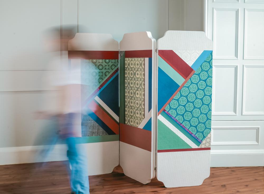 A dynamic image of a room divider with geometric designs on it in blues, greens, dark red and white, with the shape of a moving person walking in front of it.