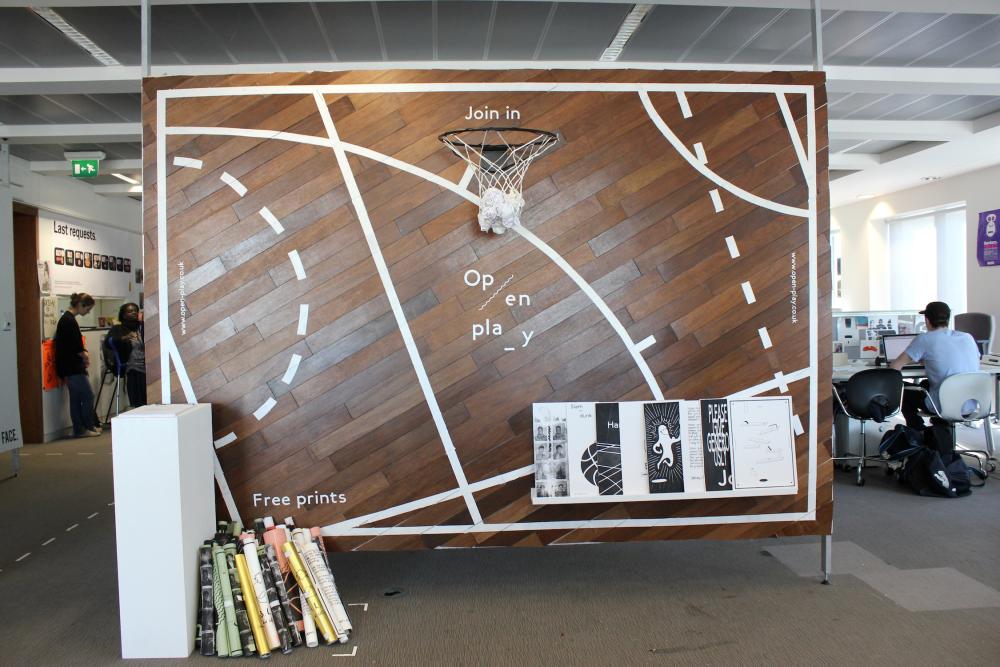 A large brown board in an office, with white lines depicting a sort of sports pitch and a basketball hoop in the upper middle, with balled up papers caught in the net. There is text that says join in, open, play.