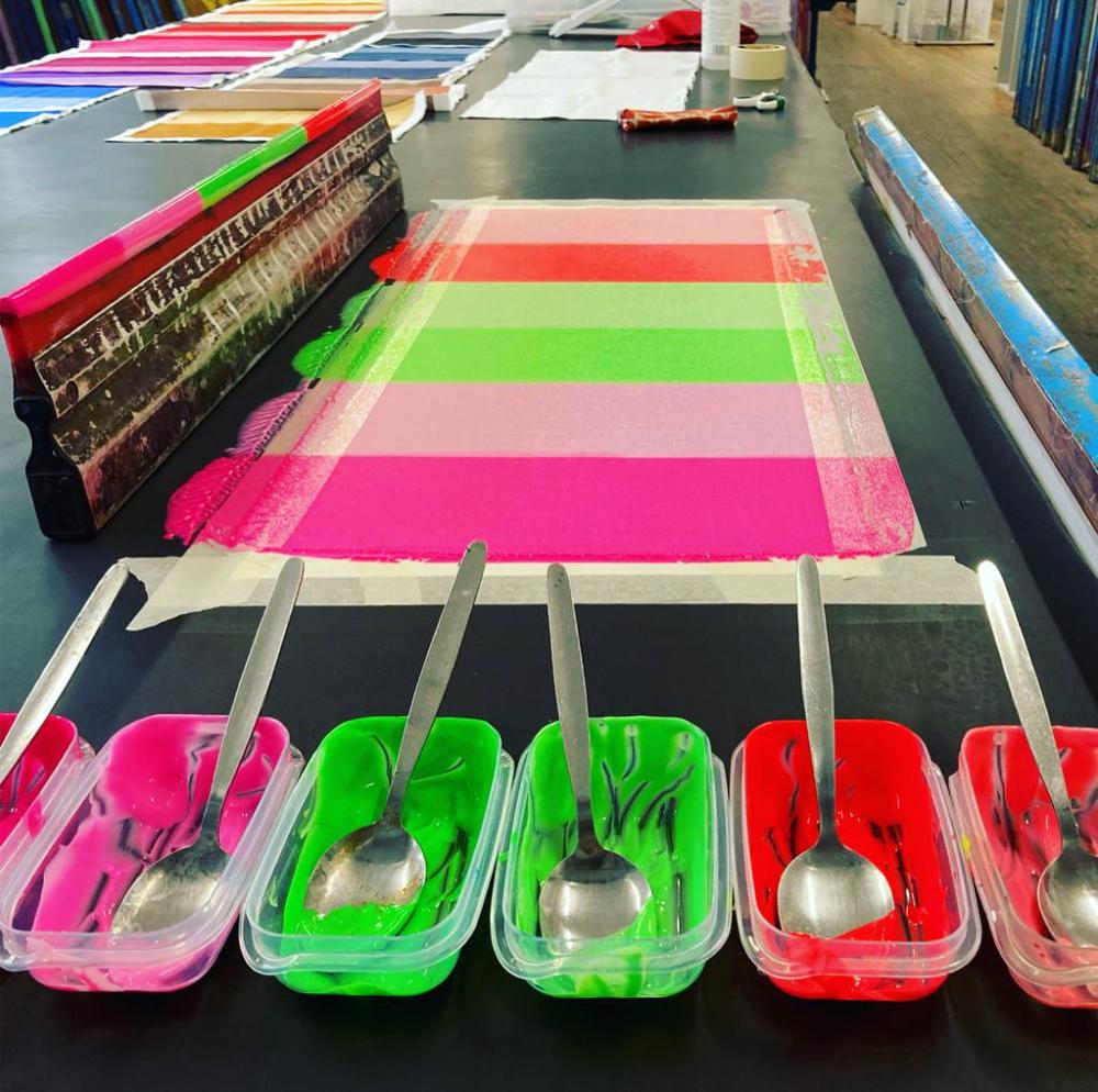 Six plastic pots of paint at the forefront of the image, each with a metal spoon, and behind them are stripes of paint painted on a desk.