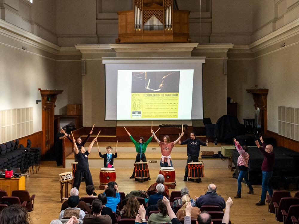 A group of people are standing behind Taiko drums in the Reid Concert Hall. They are holding drumsticks and have their arms raised.