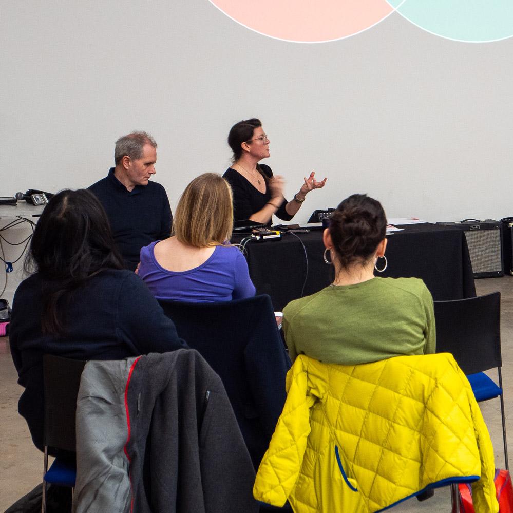 A person is seated and speaking while gesturing with their hands in front of an audience. They are listening intently.