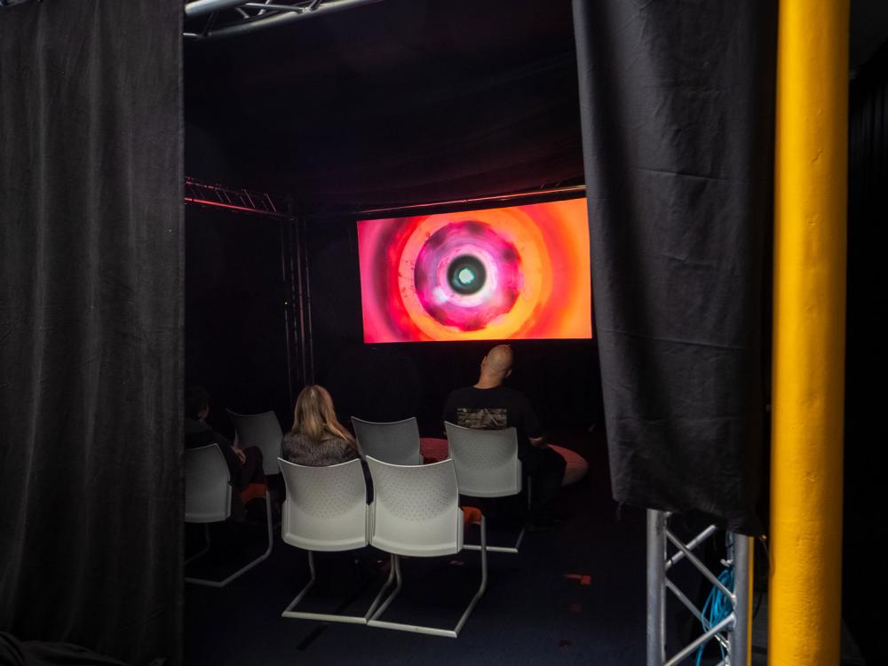 A glowing TV screen illuminates a dark room. People are sitting facing the screens.