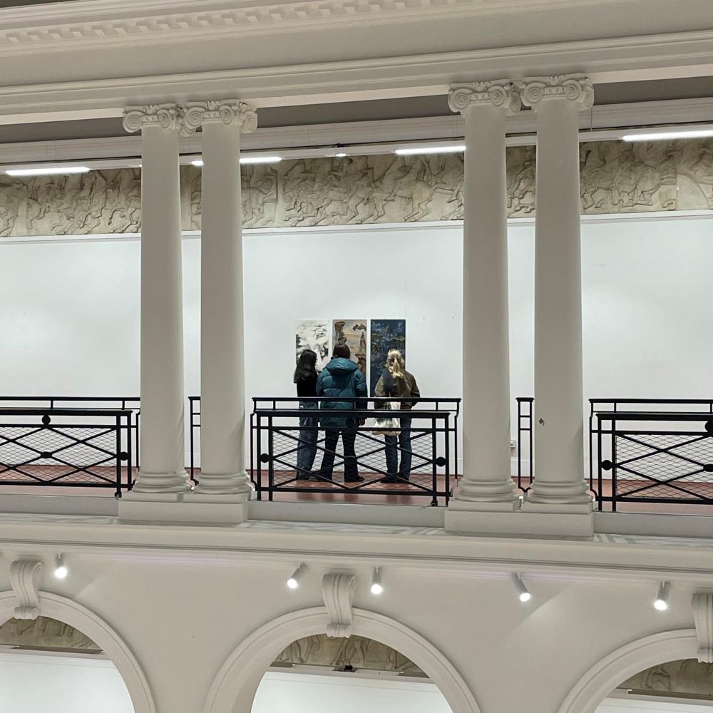 Three people viewing a piece of textiles work on the balcony of the Sculpture Court at Edinburgh College of Art. The photo is taken from the balcony across the room. 