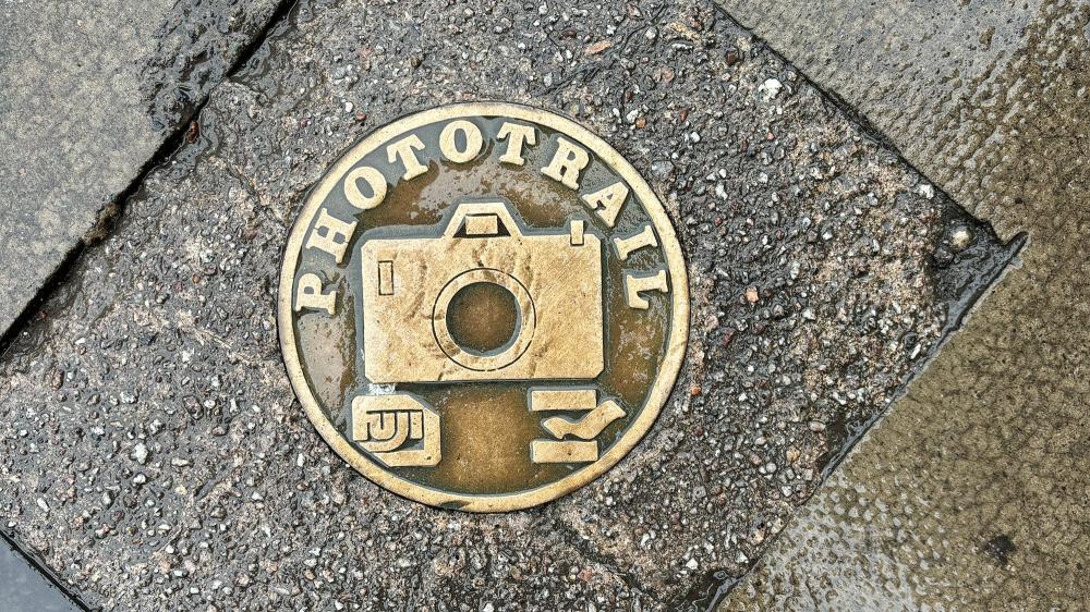 Photograph of a tile on the ground which represents the Fuji Phototrail, with a camera in the centre.