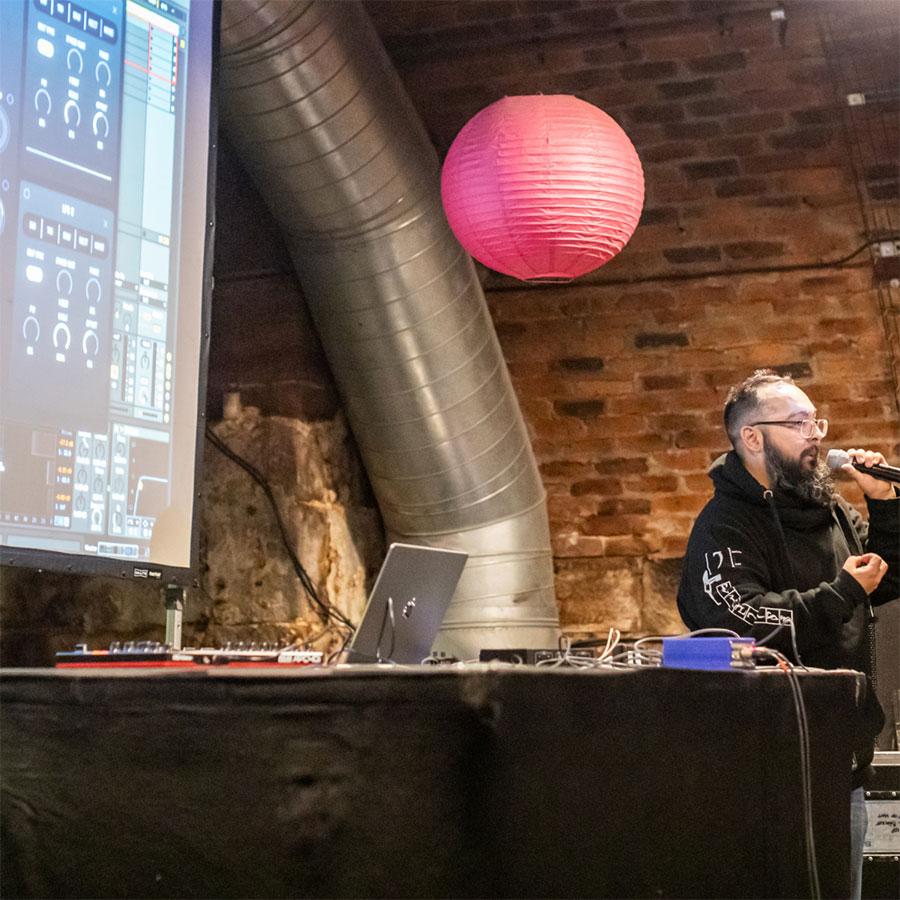 A person in a black hoody, holding a microphone and standing in a arched room, in front of a projector screen.