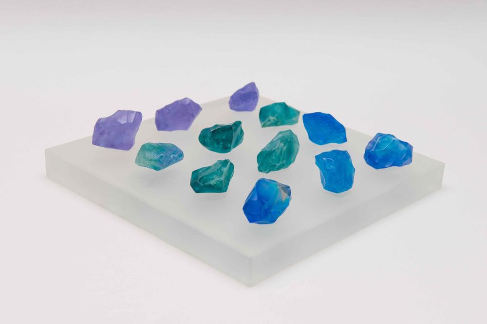 A square glass tile with small chunks of blue, turquoise and purple glass upon it.