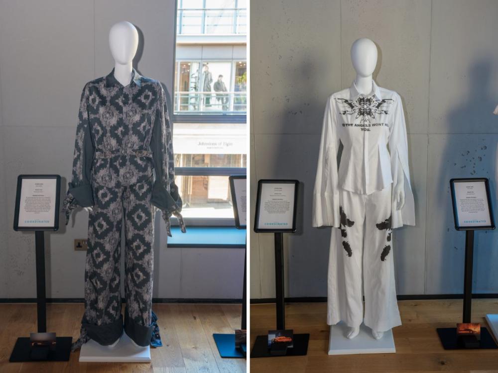 Two sets of garments on mannequins, each with trousers and a top. One is a dark grey with with a lighter pattern; the other is white with some black printing on.
