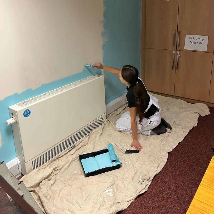 A student wearing white overalls is seated on a dust sheet on the floor. They are painting pale blue paint on a wall with a brush.