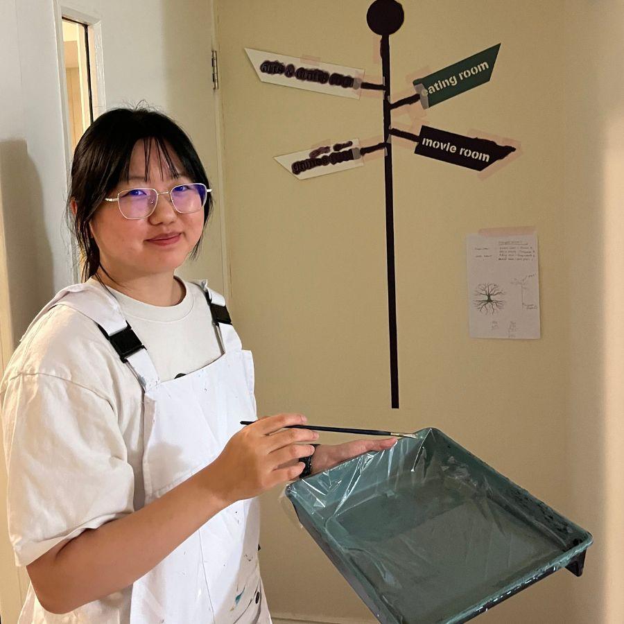 A smiling student wearing white painting overalls is smiling at the camera. They are holding a paint tray and thin brush.