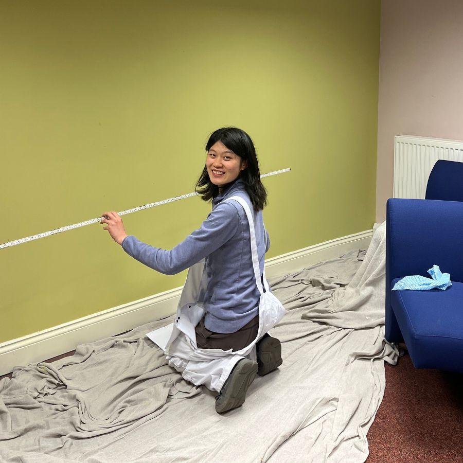 A student wearing painting overalls is kneeling on the floor and holding a measuring stick. She is smiling as she looks at the camera.