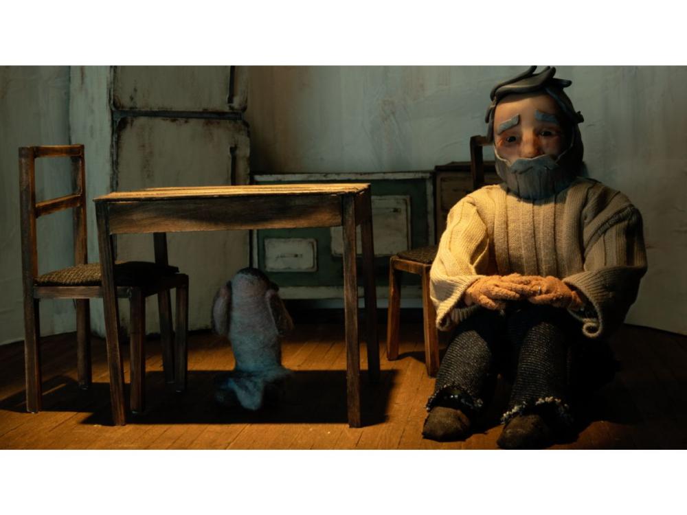The lighthouse keeper – a puppet – sits at a chair by a bare table, in a bare room. He has a grey beard and hair, and bushy eyebrows. He wears a chunky white jumper. His hands are folded on his lap and he gazes into space.