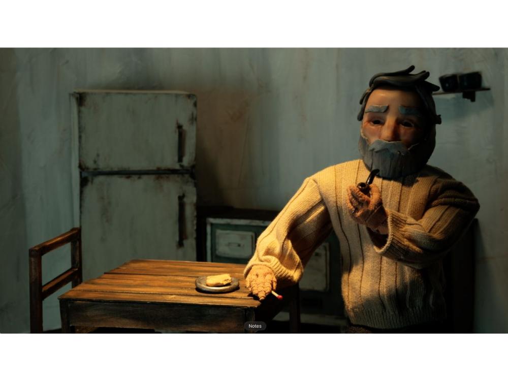 The lighthouse keeper – a puppet – sits at a chair by a bare table. He has a grey beard and hair, and bushy eyebrows. He wears a chunky white jumper. He is smoking a pipe with his left hand, a match in his right hand resting on the table. On the table is a small plate with food on it – perhaps a slice of bread – with a bite out of the corner. On the wall behind we see a shabby-looking fridge freezer or cupboard, a drawer in the corner of a dresser, and a small, high shelf.
