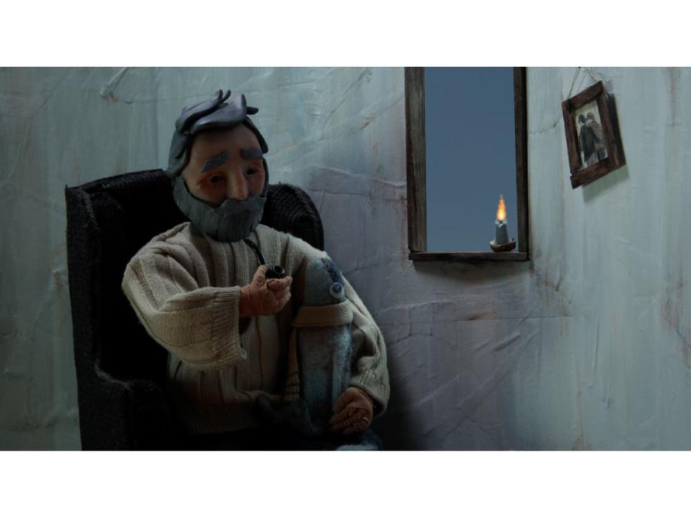 The lighthouse keeper – a puppet – sits at a chair by a window, with a candle on the window ledge. A small picture of two people side by side hangs from the wall. He has a grey beard and hair, and bushy eyebrows. He wears a chunky white jumper. He is smoking a pipe.
