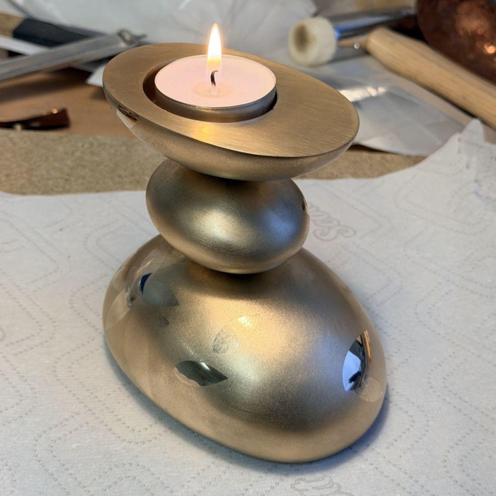 Three pebble-like metal shapes are stacked on top of each other. The top one is just the bottom half of the pebble shape, with a flat top surface, a lit candle sits in a recess at the top.