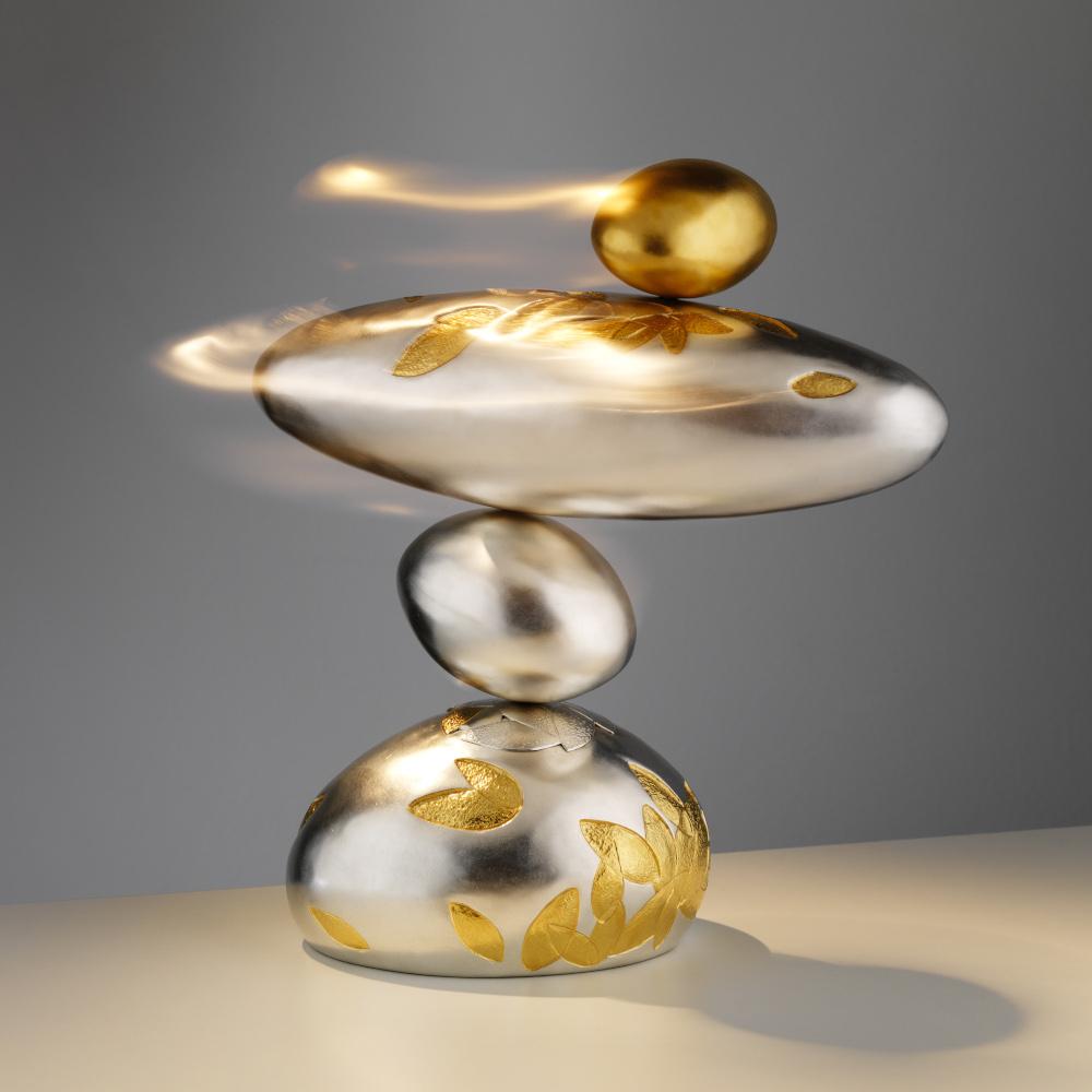 Four pebble-like metal shapes in silver and gold coloured metal, sit atop each other. Lines suggest movement, as if they're spinning around, while balanced on each other.