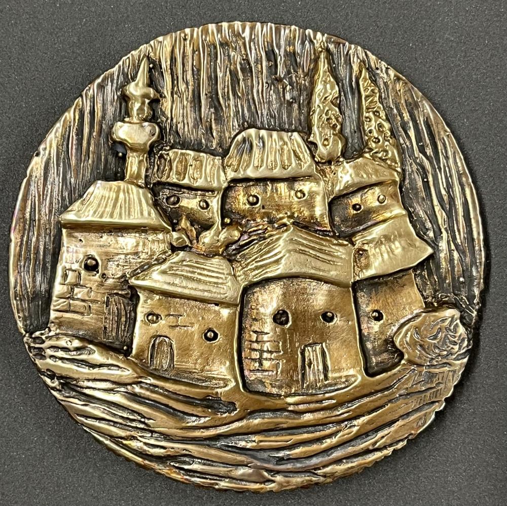 A circular medal in gold and black colouring. Small houses and buildings crowd together, two tall cypress trees reach for the sky, along with a minaret.