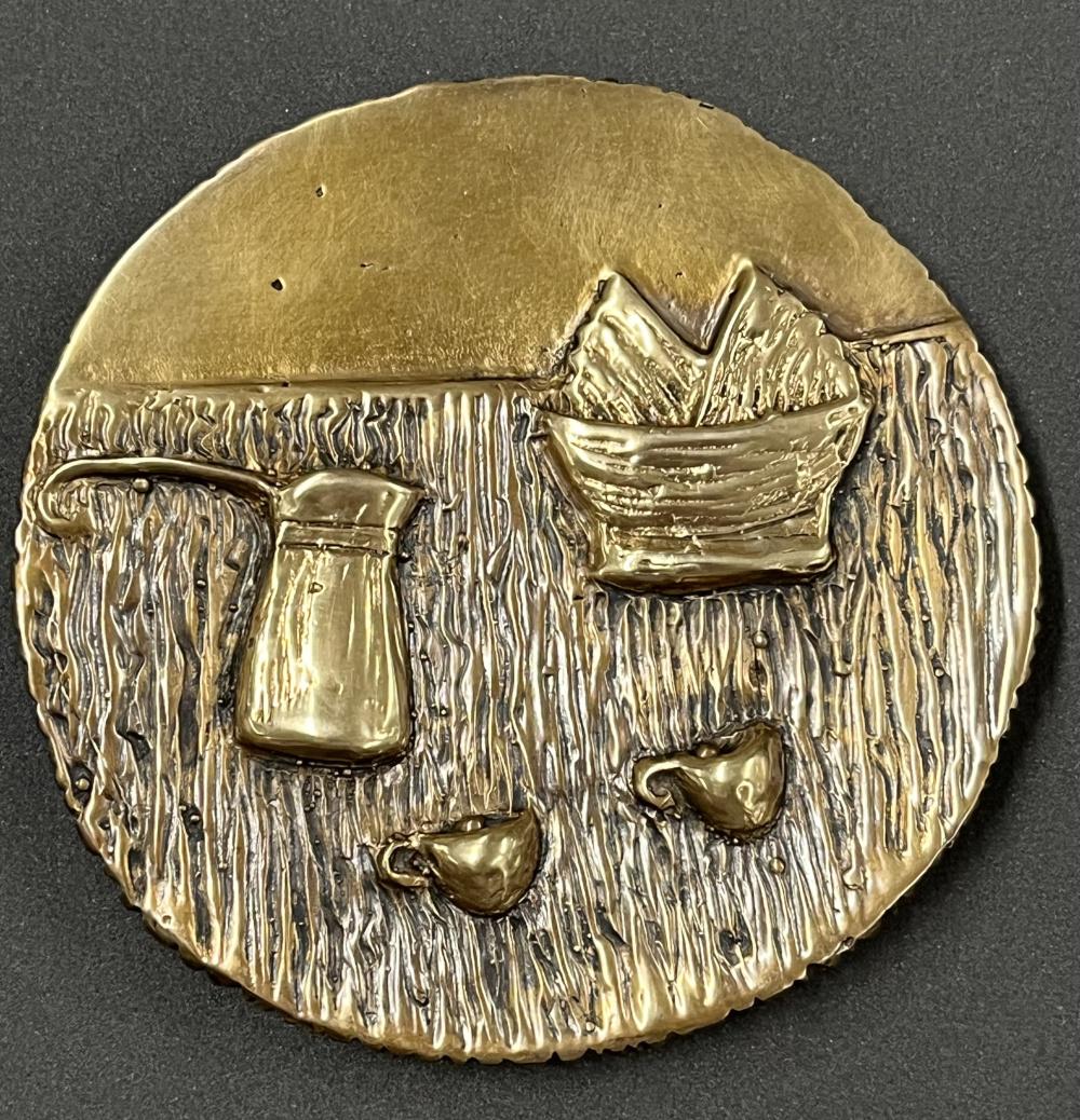 A circular medal in gold and black colouring, depicting a coffee pot two small coffee cups, and a basket with two square items in it.
