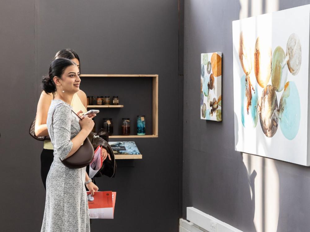 Visitors to the Graduate Show view Carol Jourdan's work - two artworks on the wall featuring abstract bubbles of colour bleeding into each other, pale blues, browns and oranges on a white background; there's a shelf on the wall holding glass bottles and jars of hand-made pigments.