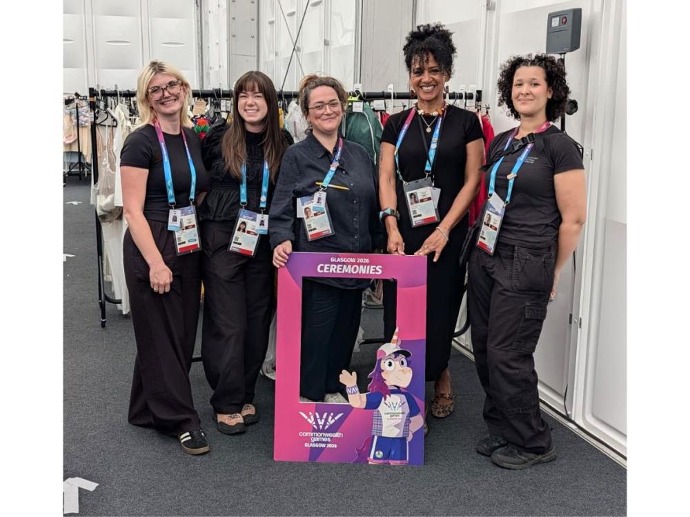 Five members of the costume team stand in a line. They're dressed in black, smiling at the camera, wearing their passes. Behind them are rails of costumes on hangers, and they hold a pink frame with Finnie, the Commowealth Games mascot on.