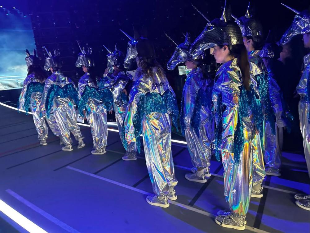 A group of performers wearing silver unicorn costumes, on stage, lit with bluish light.