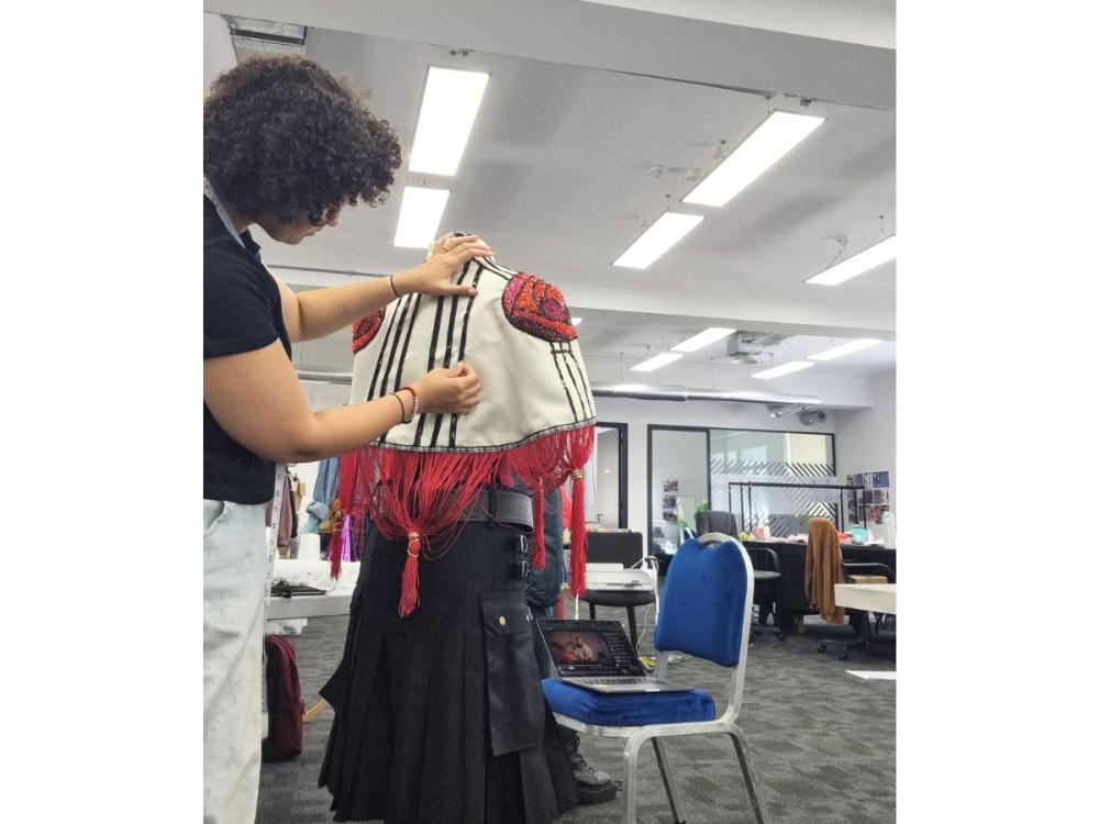 Izzy works on a piece of costume - it is a short, mid-back length cloak in cream with black lines, red tassles, and a red beaded rose on each shoulder, in the style of Rennie Mackintosh