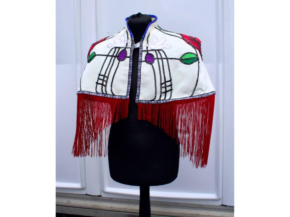 On a dressmaking form, a short, mid-back length cloak in cream with black lines, red tassles, a red beaded rose on each shoulder, green leaves and purple petals, in the style of Rennie Mackintosh