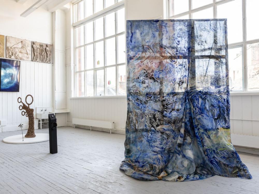 A studio at the Graduate Show. In the foreground is a large, mounted piece of fabric with blue swirling patterns on it; in the background are some sculptures, including one that looks a little like a tree, the branches forming circles.