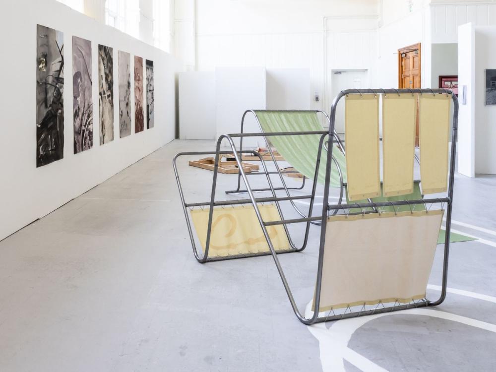 A studio at the Graduate Show. There are photographs on the left-hand wall. On the floor are several structures of curved metal and canvas, looking like playground equipment or sun loungers.