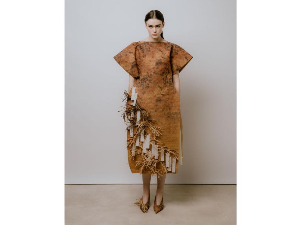 A model wears a brown dress with a cork-like texture and a print a little like a smudged animal print. Hanging from it are white rectangles and bunches of straw-like material.
