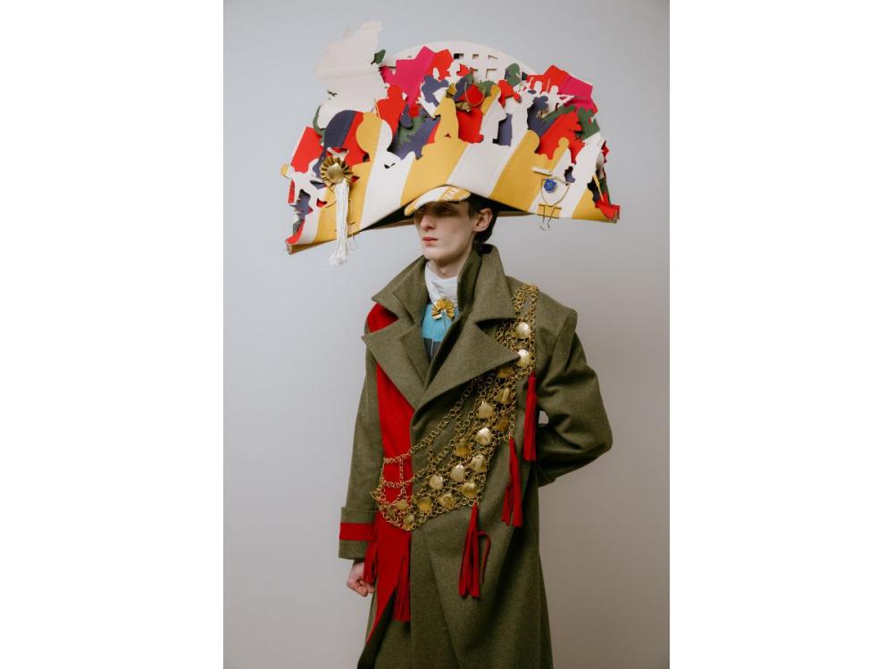A model wears a huge, elaborate hat made of multiple outlines of people in different colours. He wears a green military-style coat with a sash made of gold-coloured chains and shields, red tassels and a red scarf.