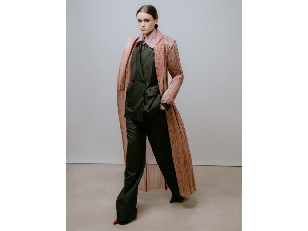 A model wears a dark coloured double-breasted suit. Over it is a long, sheer overcoat with swirling lines of pinkish colour running the length of it.
