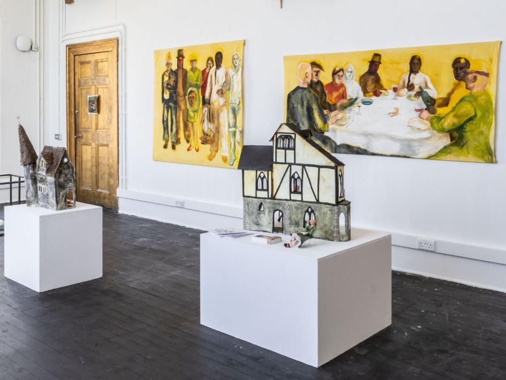 A studio at the Graduate Show. On the rear wall are two paintings of groups of people gathered together. In the foreground are sculptures of houses on white plinths.