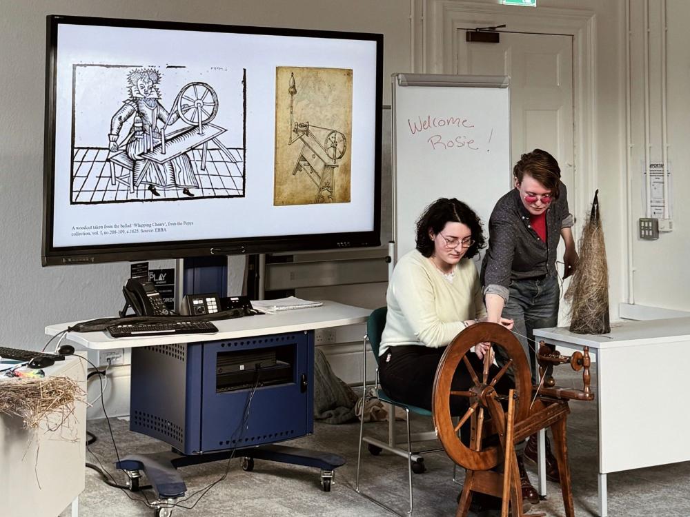 Two people experiment with using a traditional spinning wheel in a classroom