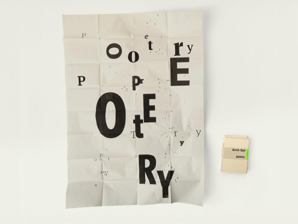 A large crumpled black and white poster covered in the repeated, exploded word "poetry" at mixed sizes, photographed flat beside a small folded zine showing a beige cover reading "dumb God poems" and a bright green spine.