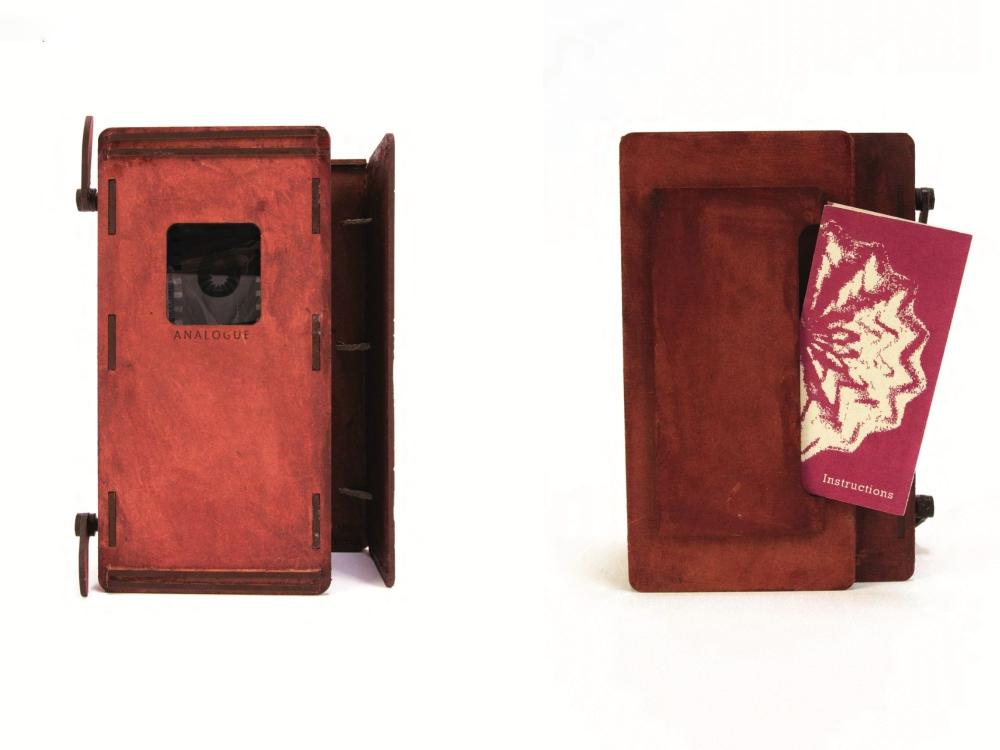 Two views of a handmade reddish-brown box object against a white background. On the left it stands upright, its front panel a mottled deep red with a small window revealing a strip of film and the word "Analogue" beneath it. On the right the box is shown from another angle, deep burgundy and wood-toned, with a small maroon booklet titled "Instructions" tucked into a recess.