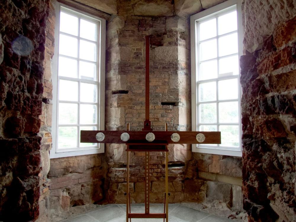 A room of bare brick and stone, with two tall windows. in the centre stands an easel, bearing a plank of dark coloured wood, with five circular pencil drawings on it. They are too small to see the detail, but are close up images of bats.
