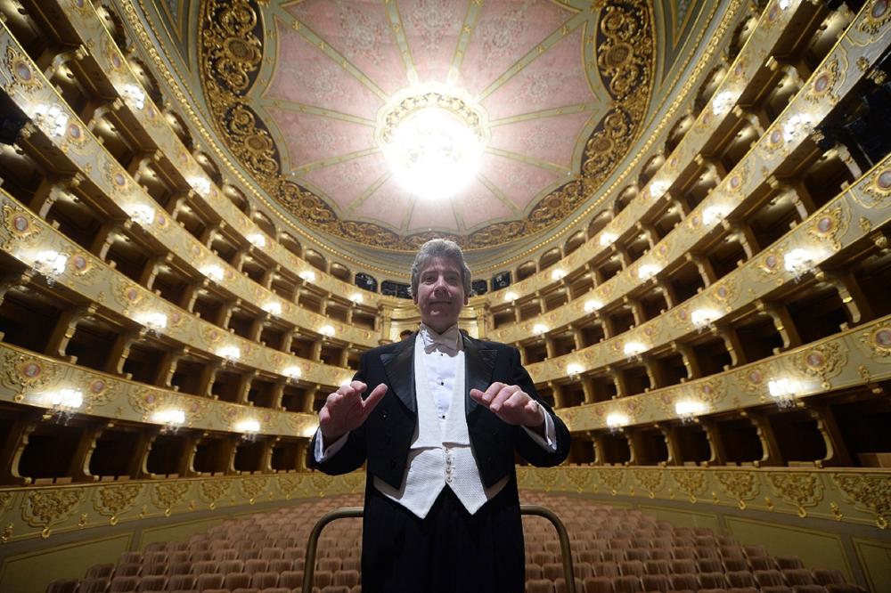 Emre Araci looks down to the camera, in a conducting pose, dressed in formal tie and tails, whilst the gilded gold and red backdrop of the Sao Carlos Opera House curves around behind him.