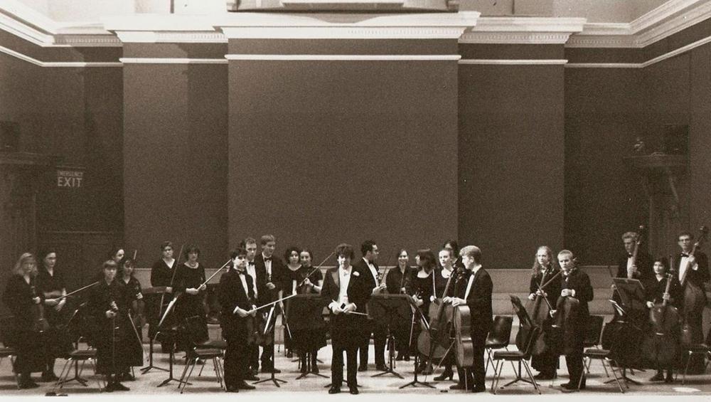 Emre Araci stands in t he Reid Concert Hall, surrounded by other members of the university string orchestra. They are all holding their instrument.