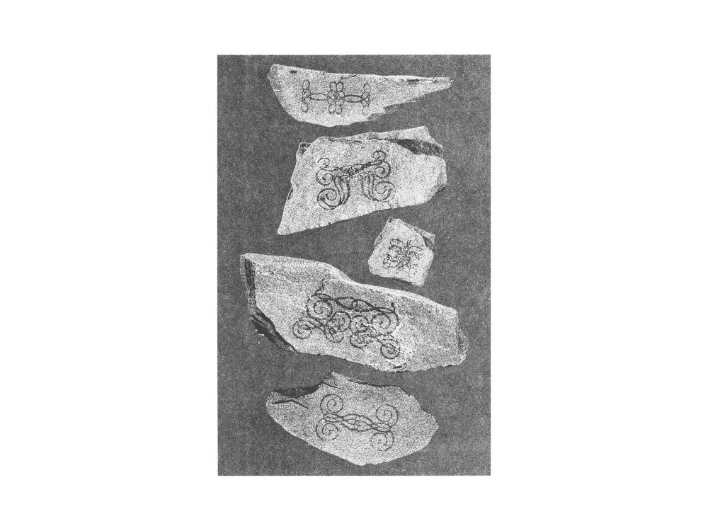 A grainy, high-contrast black and white photograph of five broken stone fragments on a dark ground, each carved with ornate looping and spiral flourishes.