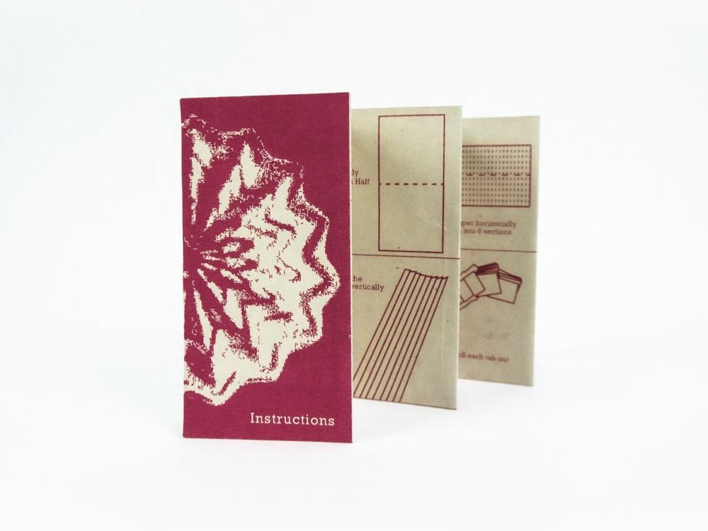 A small booklet stood upright on a white background, printed in deep burgundy and cream. The front panel carries a textured illustration and the title "Instructions"; the fanned-out inner panels show diagrams and step-by-step directions for folding and cutting paper, including "fold in half", "cut vertically", "into 8 sections" and "pull each tab out".